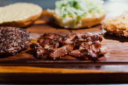 A plate of bacon and a hamburger on a wooden cutting board. The bacon is sizzling and the hamburger is to the left of the baconの写真素材