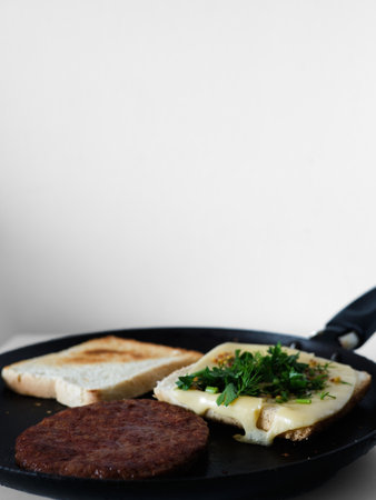 A black pan with a hamburger, cheese, and greens on top. The hamburger is cooked and the cheese is...