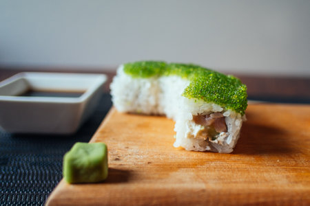 A sushi roll with green seaweed on top sits on a wooden cutting board. A small green cube sits next to the sushi rollの写真素材