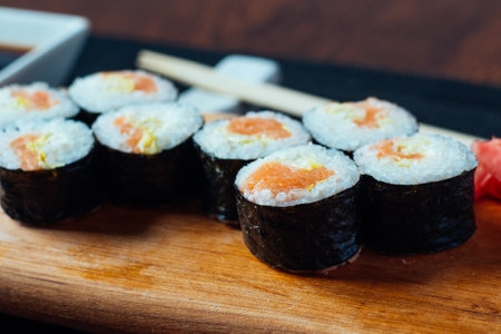 A sushi roll with a piece of ginger on top. The sushi is on a wooden board. The sushi roll is surrounded by chopsticksの写真素材