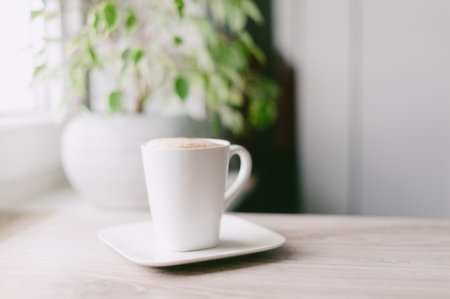 A white coffee cup with a white saucer sits on a wooden table. The cup is filled with coffee and has a plant in the backgroundの写真素材
