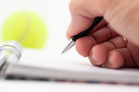 A person is writing in a notebook with a pen. A tennis ball is sitting on the table next to the notebookの写真素材