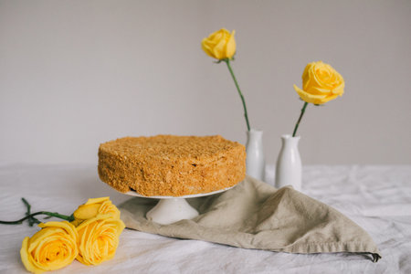 A cake sits on a white plate on a table with a yellow cloth draped over it. A vase of yellow roses sits next to the cake. Concept of warmth and happiness, as the bright yellow cakeの写真素材