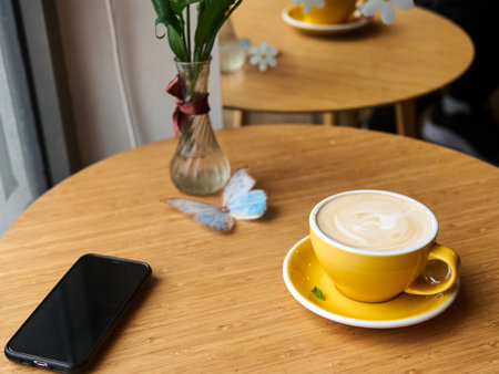 Yellow coffee cup sits on a table next to a cell phone. The cup is filled with coffee and has a white design on it. The table is made of wood and has a vase with flowers on it. The scene is cozyの写真素材