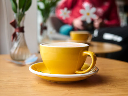 Yellow coffee cup sits on a yellow saucer on a wooden table. The cup is filled with coffee and has a white foam on top. The table is surrounded by vases with flowers, creating a cozyの写真素材