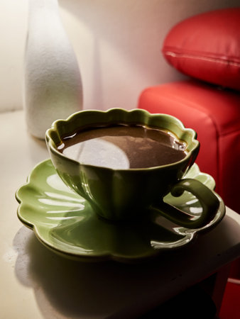 Green cup with a flower design sits on a white table. The cup is filled with a dark liquid, possibly coffee or tea. The table is surrounded by a red couch and a white vase. The scene conveys a cozyの写真素材