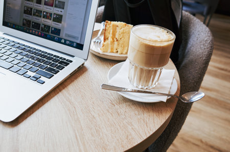 Laptop is on a table with a cake and a cup of coffee. A fork is on the cakeの写真素材