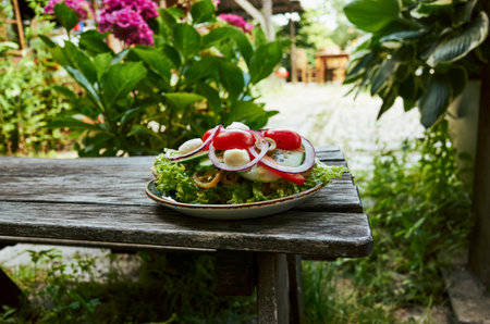 Plate of salad with tomatoes, cucumbers, and onions is sitting on a wooden table outsideの写真素材