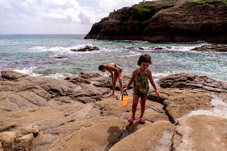 Caucasian girl playing with a bucket of water among the rocks with her motherの写真素材