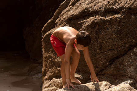 caucasian boy in red swimming trunks climbing a rock on the beach in summerの写真素材