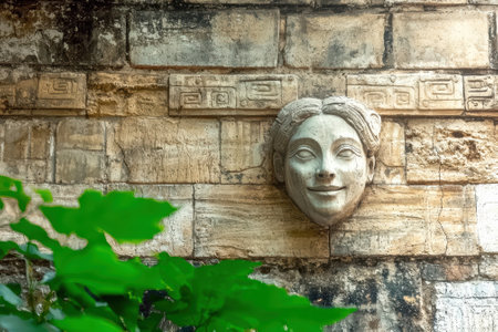 Stone wall with carved face and green leaves.の素材