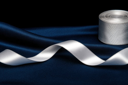 Elegant silver ribbon on dark blue satin fabric with a roll of ribbon in the background.の素材