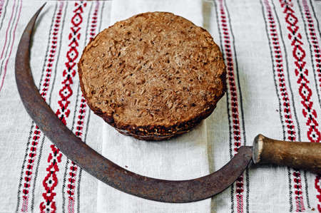 Dark round bread on woven cotton towel with traditional ornament and sickle agricultural toolの写真素材