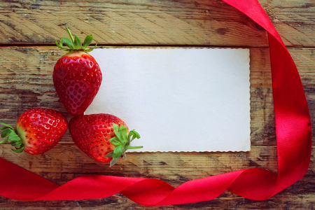 three strawberries, red satin ribbon and empty paper tag on an old wooden tableの写真素材