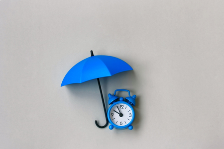 Blue alarm clock under an umbrella on a gray background, concept of protection time