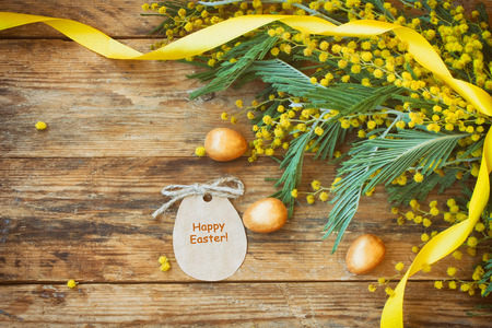 Easter holiday background with branches of mimosa, golden eggs, paper tag with the inscription "happy easter" on a wooden tableの写真素材