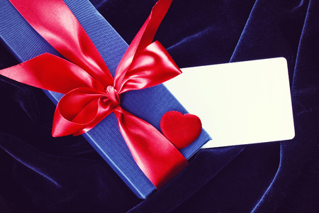 Holiday blue gift box with red ribbon, silk heart, empty paper tag on dark blue velvetの写真素材
