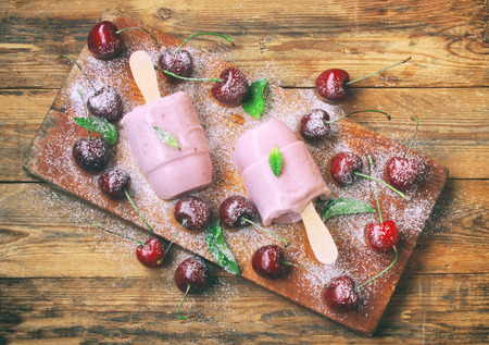 Homemade yogurt ice cream, with cherry, popsicle on stick on wooden board on table, powdered sugar, top viewの写真素材