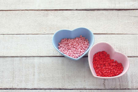 Valentines day background, many small red pink sugar hearts in bowls on white wooden table, top...