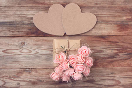 two cardboard heart, gift box wrapped in kraft paper, bouquet of pink roses on wooden table, top...
