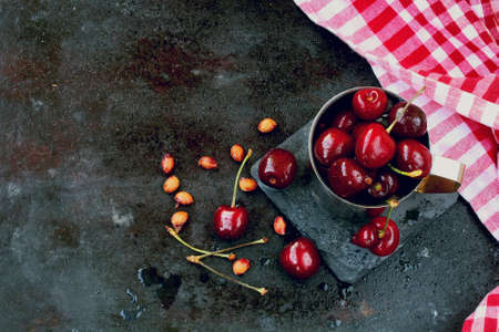 sweet ripe cherries and pits in metal mug on old black scratched background, checkered towel, top...