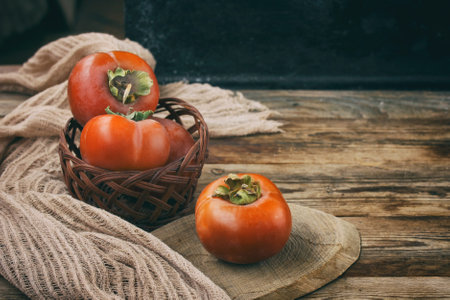 sweet ripe persimmon in wicker bowl and cloth napkin, on wooden table, rustic style, dark background