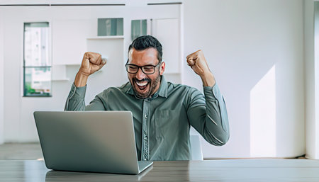 Cheerful man celebrating success while working on his laptop at homeの写真素材