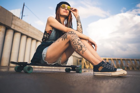 young girl with tattoos and dreadlocks in a blue cap sits on a longboard against the background of...
