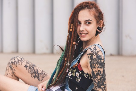 young girl with tattoo and dreadlocks is sitting on the background of concrete structureの写真素材