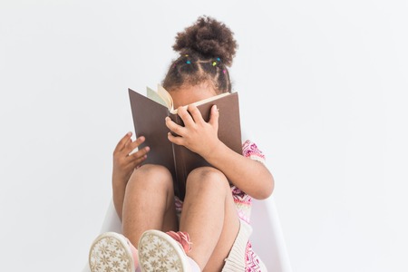 Portrait of a little girl in a colorful dress reading a book on a white backgroundの写真素材
