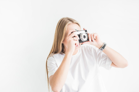 Portrait of a young stylish blonde girl using and taking pictures on an old vintage camera on a white backgroundの写真素材