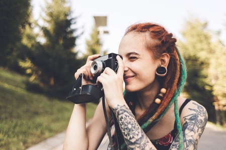 young girl with tattoos and dreadlocks photographs vintage camera in the park