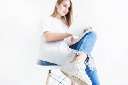 Portrait of a young stylish blonde girl in a white T-shirt and blue jeans using a tablet on a white backgroundの写真素材