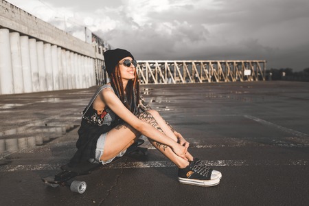 young girl with tattoos and dreadlocks in a blue cap sits on a longboard against the background of concrete structureの写真素材