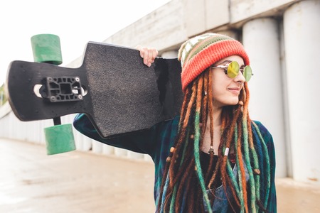 young girl in sun glasses and a cap stands with a longboard against the background of concrete...