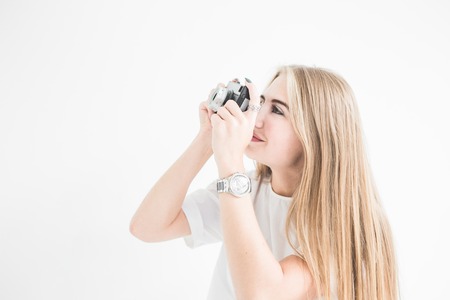 Portrait of a young stylish blonde girl using and taking pictures on an old vintage camera on a white backgroundの写真素材