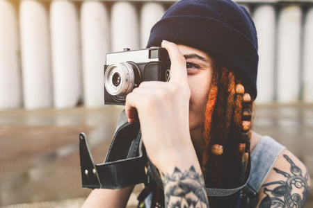 young girl with tattoos and dreadlocks in a blue hat photographs a vintage camera on the background of a concrete wallの写真素材