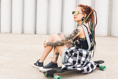 young girl with tattoo and dreadlocks sitting on a longboard on the background of concrete structureの写真素材