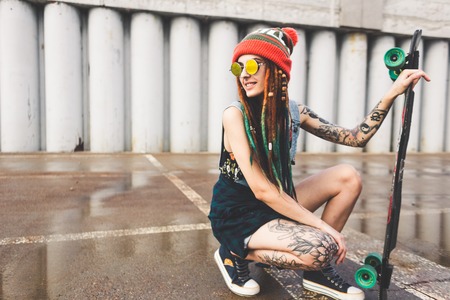 young girl with tattoos and dreadlocks in a cap sits with a longboard on the background of concrete structureの写真素材
