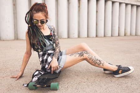 young girl with tattoo and dreadlocks sitting on a longboard on the background of concrete structureの写真素材
