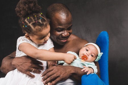 Muscular African American man sitting with children in a blue chair against a black wallの写真素材