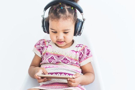 Cute little girl in a multi-colored dress, listens to music with headphones and uses a smartphone on...
