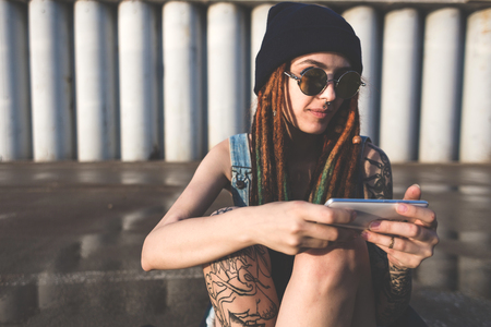 young girl with tattoos and dreadlocks in a blue cap uses a smartphone against the background of a concrete wallの写真素材