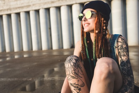 young girl with tattoos and dreadlocks in a blue cap sits on a longboard against the background of concrete structureの写真素材