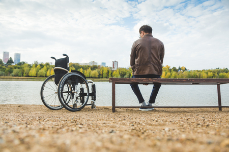 A young man sits on a bench by the lake, next to his wheelchair.の写真素材