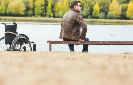 A young man sits on a bench by the lake, next to his wheelchair.