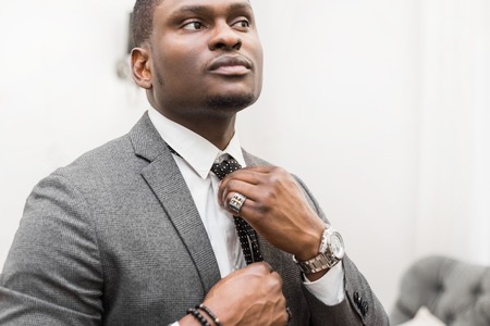 Young African American businessman in a gray suit tying a tie on a white background.