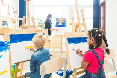 A group of preschool students and young teacher in drawing class paint in gouache. mixed race...