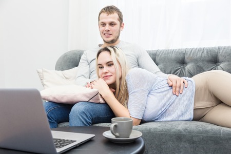 Young beautiful couple resting on the couch and watching online video from a laptop in the living room.の写真素材