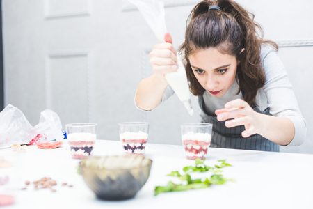 A confectioner prepares a trifle in three cups. Desserts are on the white table in the kitchen. The concept of homemade pastry, cooking cakes.の写真素材